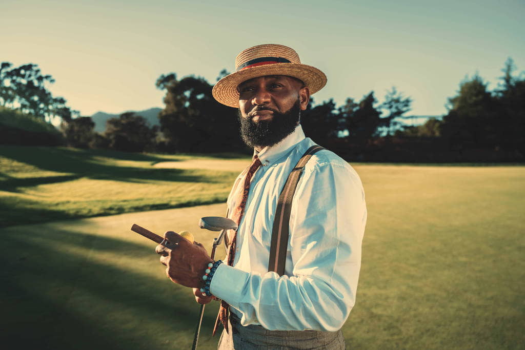 A portrait of a dashing mature bearded black man in a fashionable outfit with a straw hat and braces standing on a golf field in the evening with a sunset, holding a golf club and a cigar in the hands